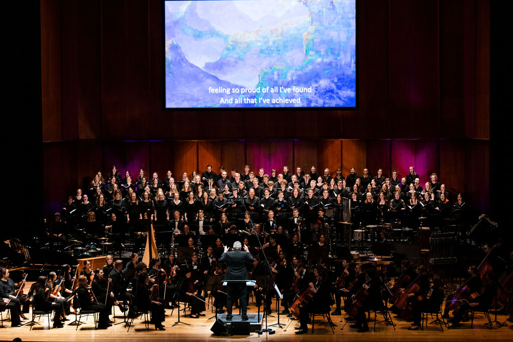 Orchestra on stage facing audience with a screen projecting images in the background.